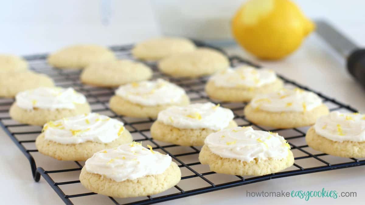 lemon drop cookies frosted with lemon icing on a cooling rack in front of a bowl of lemon icing and a zested lemon